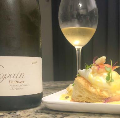A bottle of Copain 2018 DuPratt Chardonnay standing next to a glass of the wine, and a crab eggs benedict.